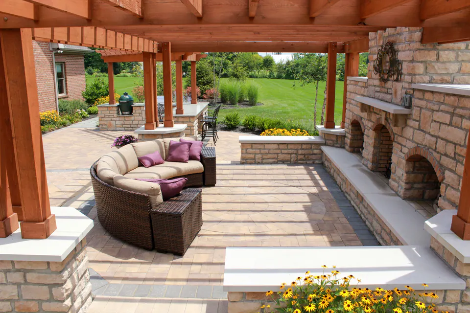 View from under a wood pergola toward stone fireplace, paver patio, lounge seating, grill counter, lawn, and planting.